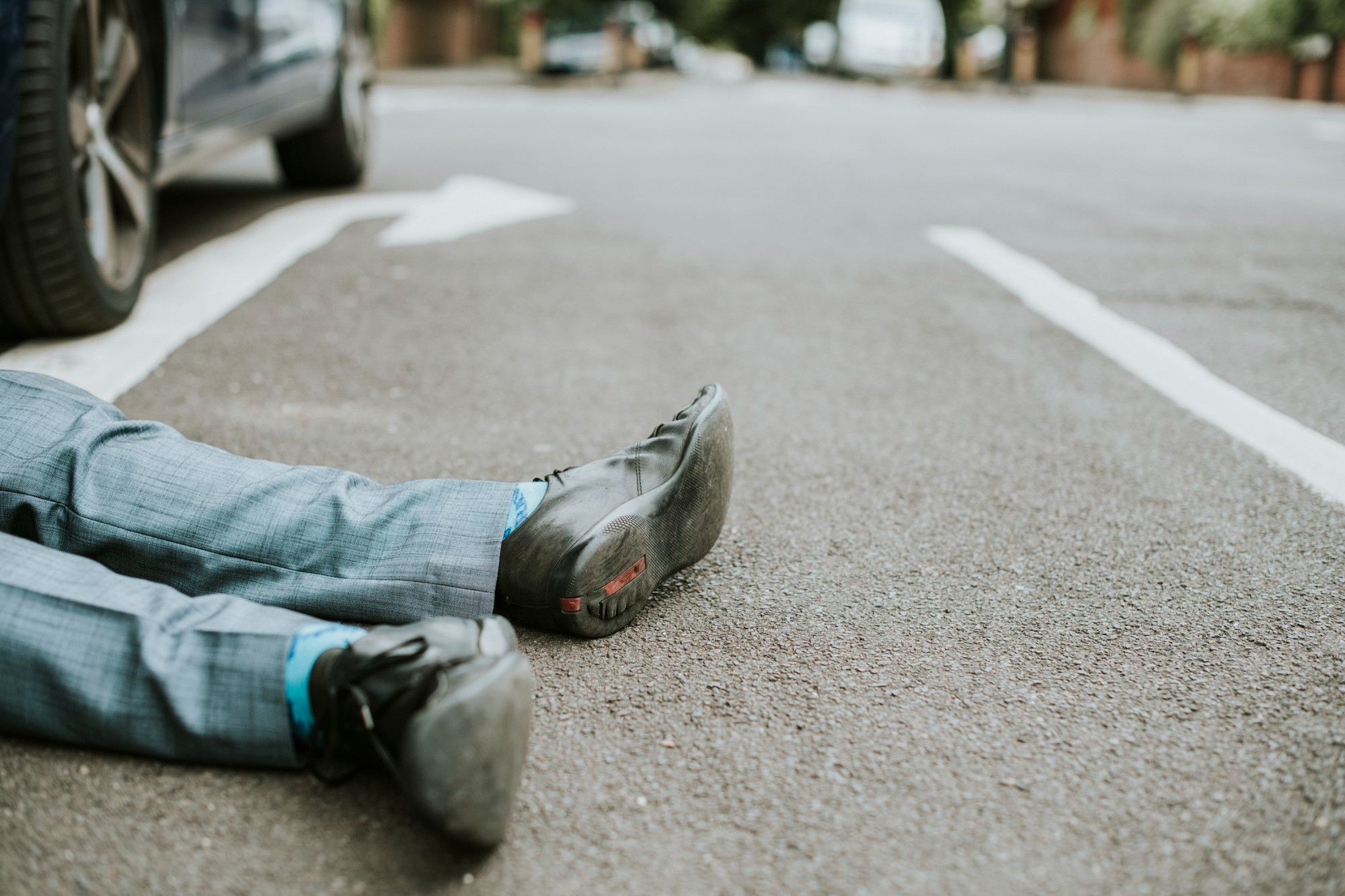 A person wearing a gray suit and black shoes, possibly a pedestrian hit by car, is lying on the asphalt of a road, with only their legs visible. A parked car and white road markings are nearby.