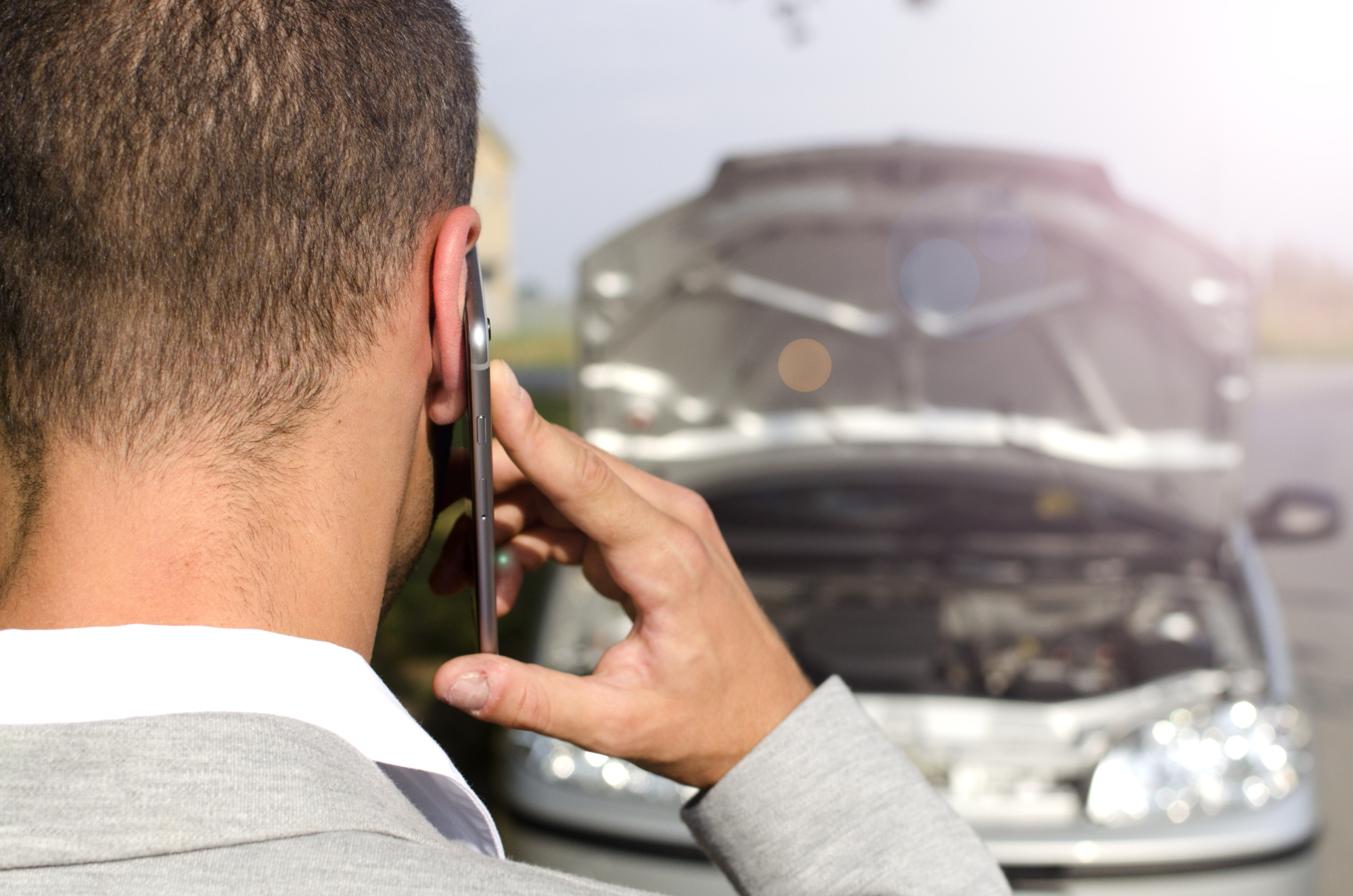 A man in a suit is seen from behind, holding a phone to his ear, standing before a car with its hood open—suggesting a possible breakdown or seeking Car Accident Injury Compensation NSW. Man standing by a broken vehicle calling someone to assist in fixing the car accident
