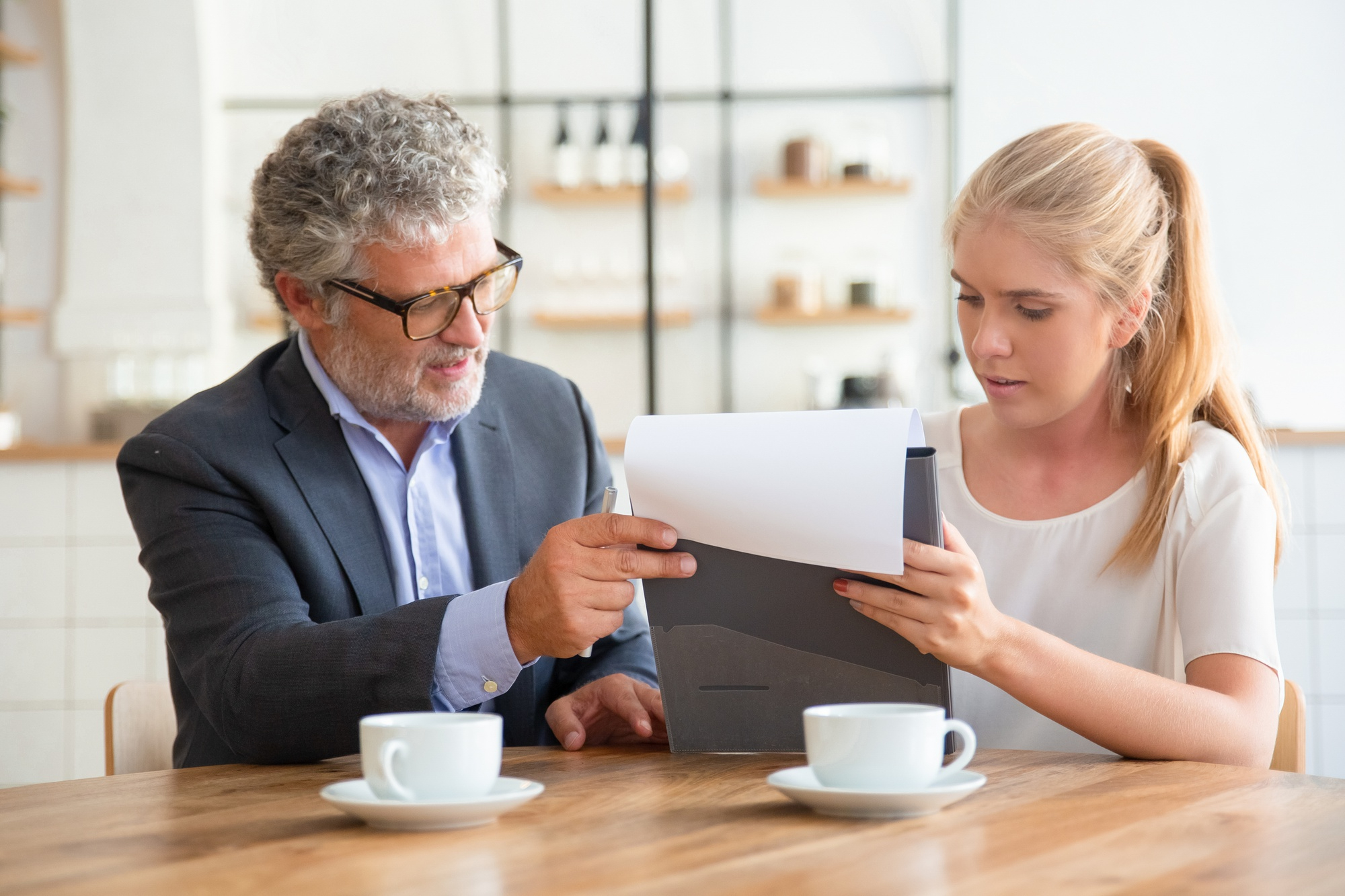 An older man in a suit and glasses sits at a table with a young woman, pointing at documents she is holding. Both are focused on the papers—likely discussing wills and estate matters. Two coffee cups are on the table in front of them.