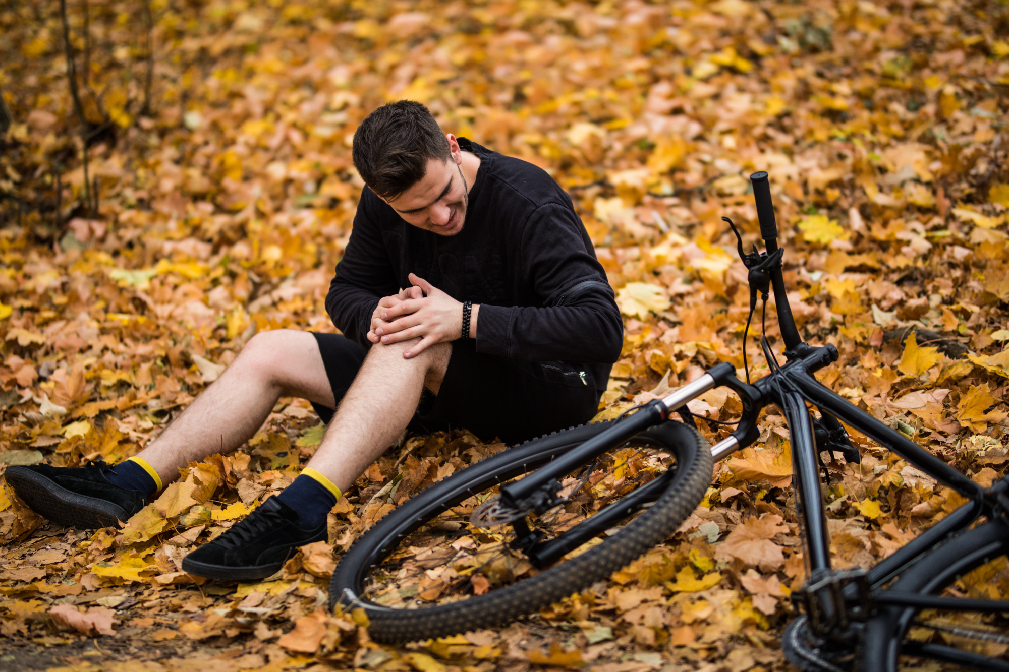 A man sits on the ground covered with autumn leaves, holding his knee in pain next to a fallen bicycle—a typical scene of cyclist accidents.