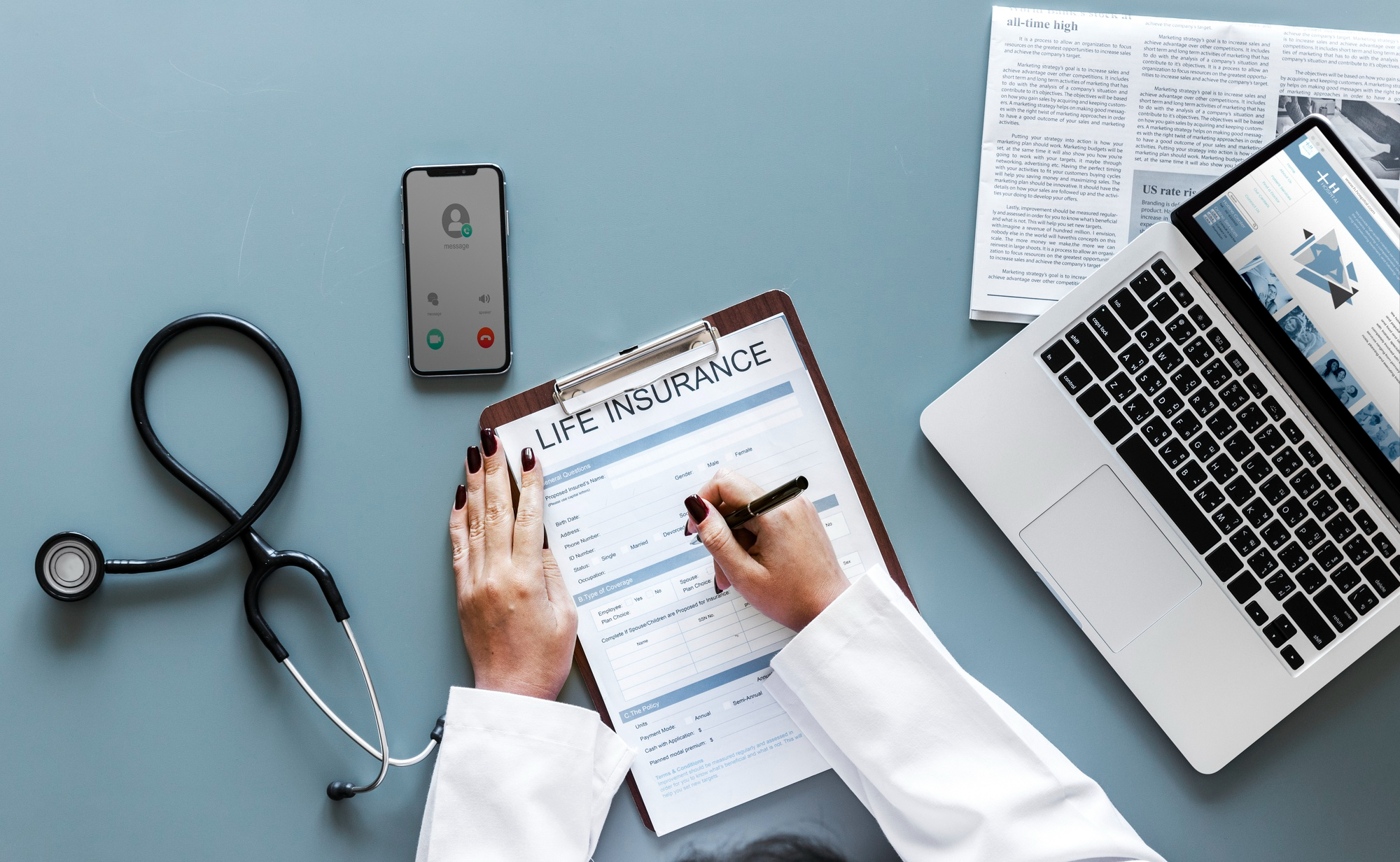 A person in a white coat fills out a life insurance form on a clipboard, with documents about head trauma, a laptop, stethoscope, and a smartphone with an incoming call on a gray desk. A person in a white coat fills out a life insurance form on a clipboard, with documents about head trauma, a laptop, stethoscope, and a smartphone with an incoming call on a gray desk.