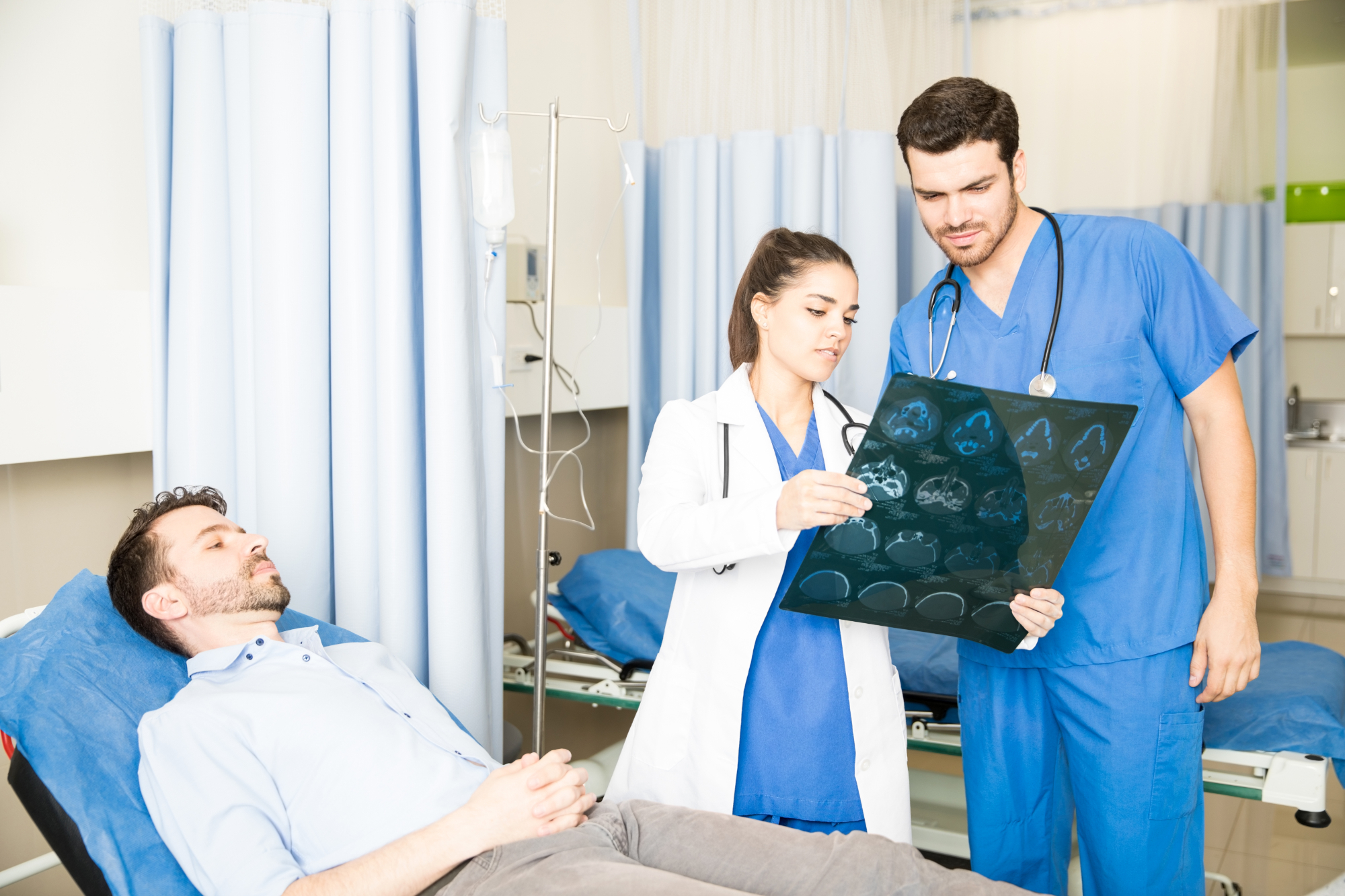 A female doctor and a male nurse examine brain scans, possibly for an Accident Compensation case, while a male patient rests on a hospital bed nearby in a medical room with blue curtains and equipment.