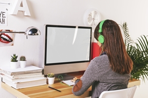 A person with long hair wearing green headphones sits at a desk, working on a computer with a blank screen—perhaps researching Total and Permanent Disability. The desk has stacked papers, cacti, and a lamp, with plants and wall decor in the background.