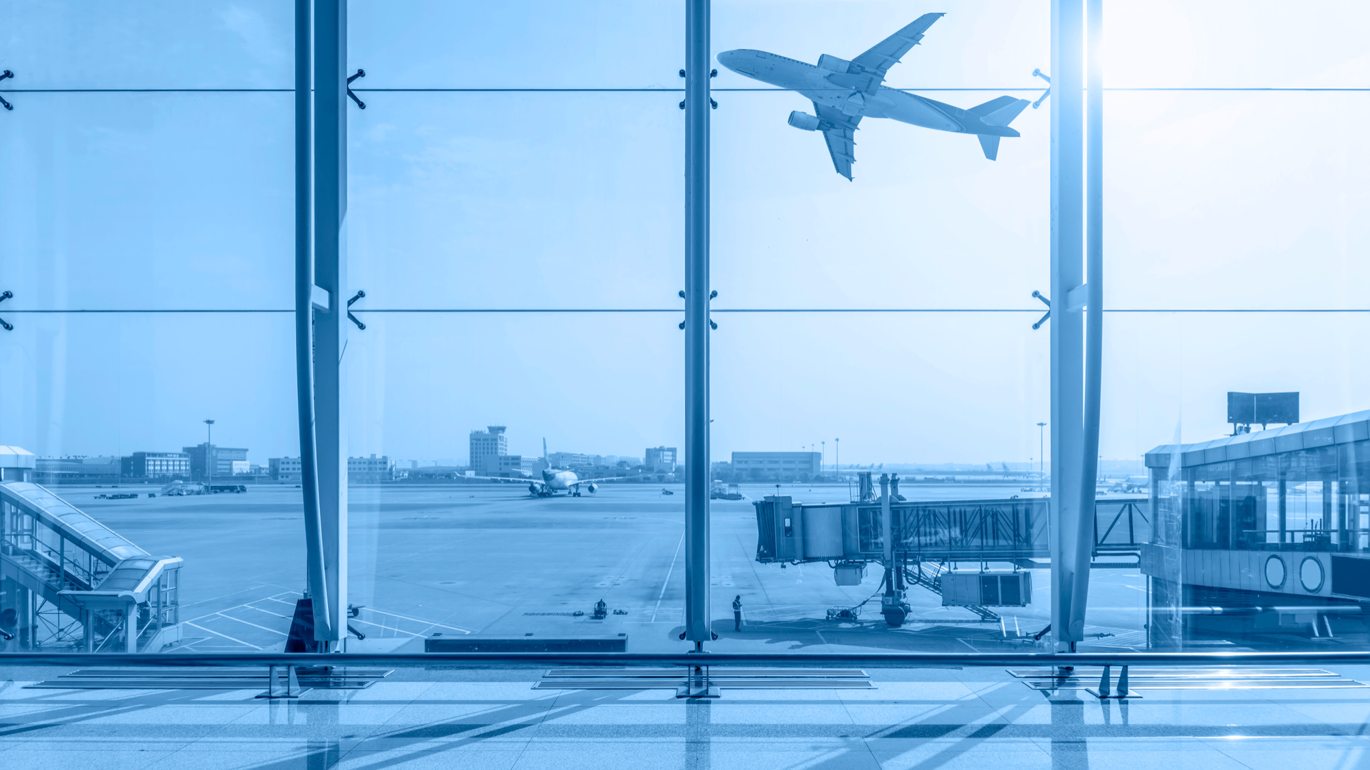 A large passenger airplane flies above the runway at an airport, viewed through tall glass windows from inside a terminal. Sunlight shines through the windows, casting reflections on the polished floor as travelers discuss compensation for delayed flights.