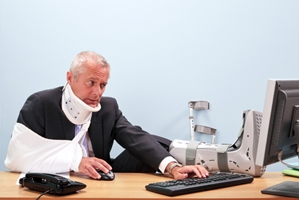 A man in a suit wearing a neck brace, arm sling, and foot cast sits at a desk using a computer, looking frustrated as he deals with workers compensation paperwork.