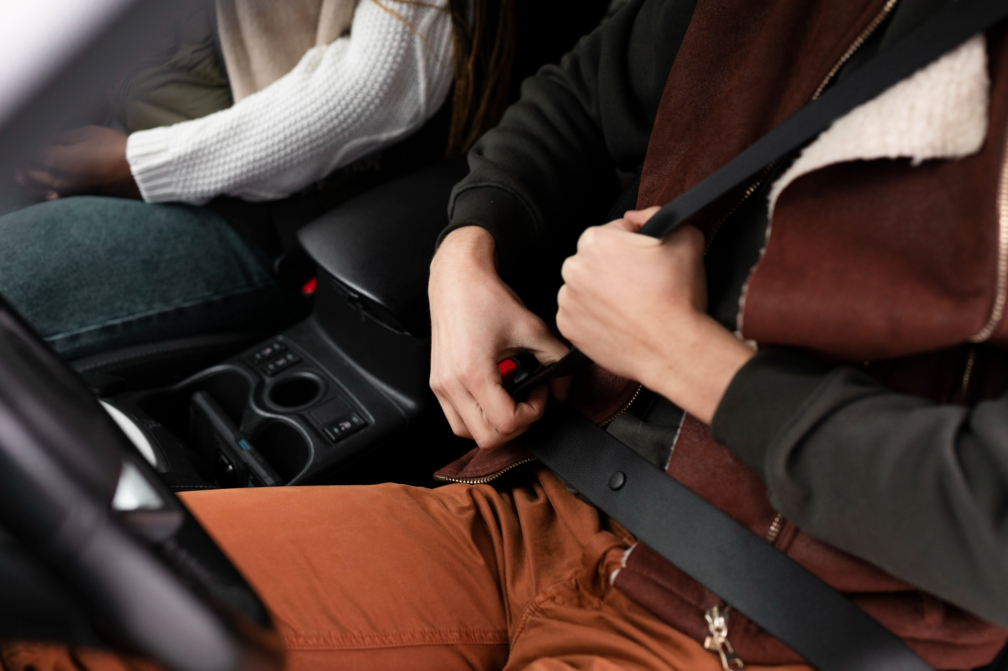 A person in a brown jacket and orange pants fastens their seatbelt in a car, sitting next to another passenger in a white sweater and blue jeans—highlighting the importance of safety to help prevent car accidents NSW.