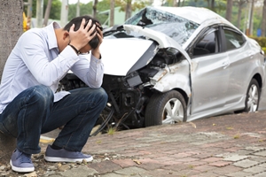 A man sits on the curb with his head in his hands, appearing distressed, in front of a heavily damaged silver car after a car accident.