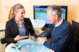 A woman in business attire shows a hearing aid device to an older man seated at a desk, discussing ways to prevent medical negligence, while a computer screen displaying data glows in the background. Both appear engaged in conversation. A woman in business attire shows a hearing aid device to an older man seated at a desk, discussing ways to prevent medical negligence, while a computer screen displaying data glows in the background. Both appear engaged in conversation.