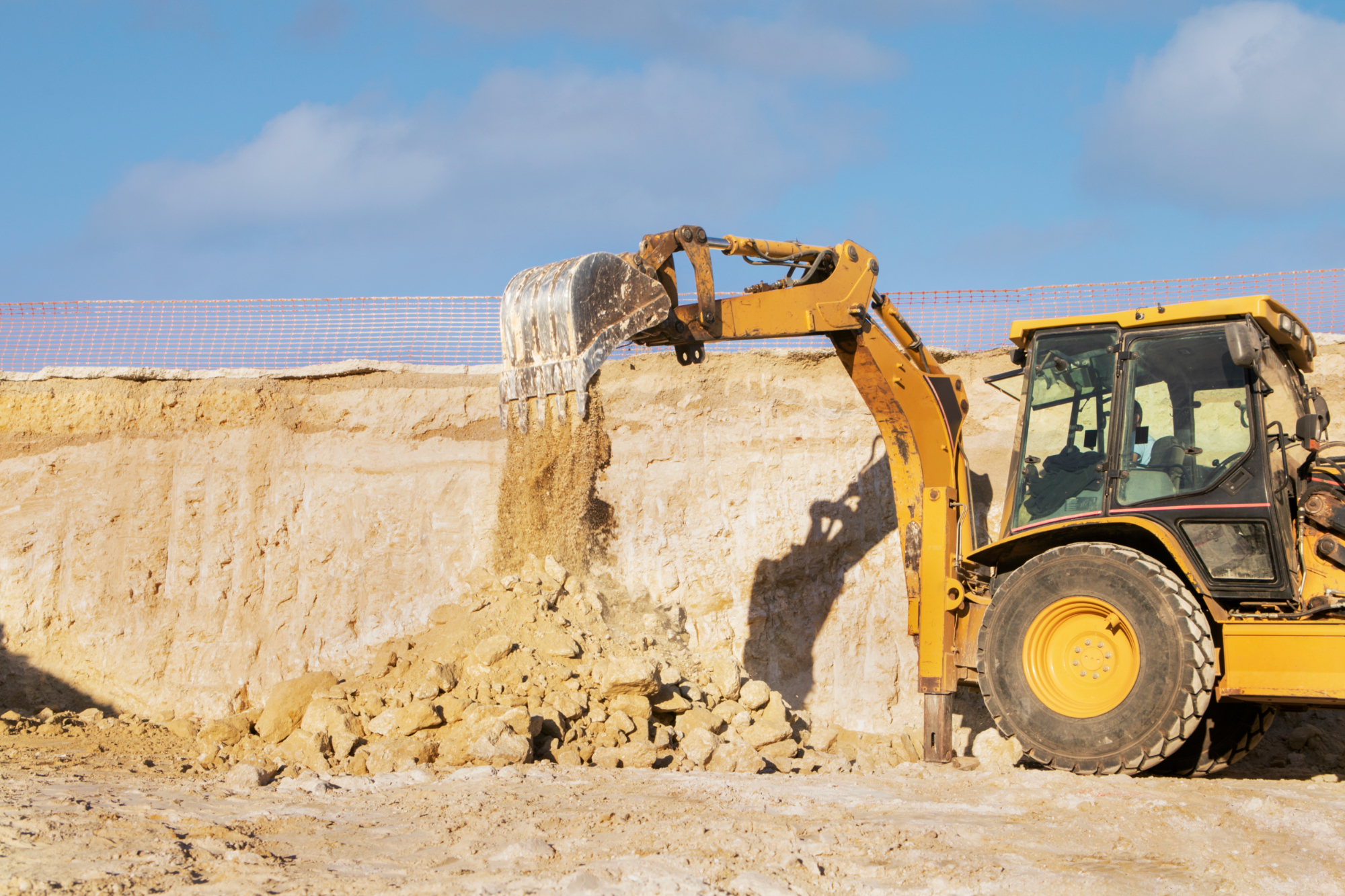 A yellow excavator dumps sand and rocks at a construction site, with a clear sky in the background and a safety fence along the edge of the excavation area, where workers are assured proper compensation for their efforts.