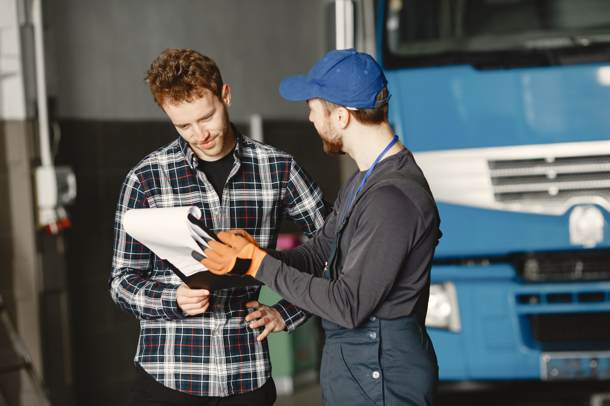 A mechanic wearing gloves and a cap shows a clipboard to a man in a plaid shirt as they discuss paperwork about accidents involving trucks, standing in front of a large blue truck inside the garage. A mechanic wearing gloves and a cap shows a clipboard to a man in a plaid shirt as they discuss paperwork about accidents involving trucks, standing in front of a large blue truck inside the garage.