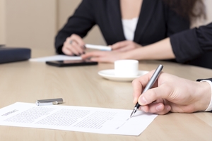 Two people sit at a desk; one is signing an injury claim document with a pen, while the other, dressed in business attire, holds a pen next to a cup and saucer.