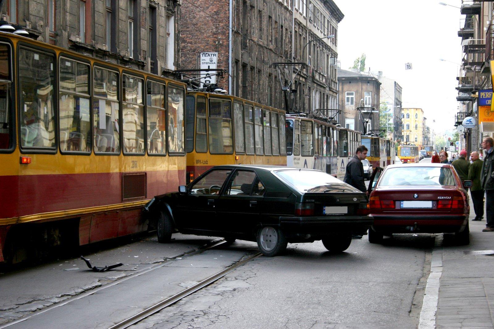 Two cars have collided on a city street, with one car crashed into the side of a yellow tram. People stand nearby observing the scene, and the street is lined with old buildings.