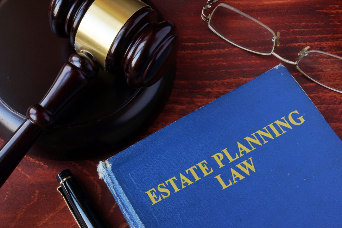 A blue book titled "Estate Planning Law" is on a wooden desk next to a gavel, eyeglasses, and a black pen.