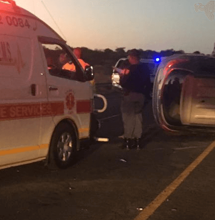 An ambulance and emergency personnel attend the scene of a car accident at dusk, with one vehicle overturned on its side on the road. Blue and red emergency lights are visible in the background.