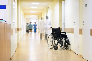 A black wheelchair sits empty in a hospital hallway, with medical staff in scrubs walking and conversing in the background. The corridor is brightly lit with white doors and pale yellow walls.
