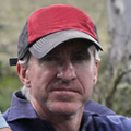 A man wearing a red and gray cap and a dark blue collared shirt looks at the camera with a neutral expression, as if considering options like injury compensation lawyers NSW. The background is blurred and appears to be outdoors.