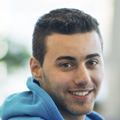 A young man with short dark hair and a trimmed beard, wearing a blue hoodie, smiles confidently at the camera. The softly blurred background hints at his positive outlook after meeting with injury compensation lawyers NSW.