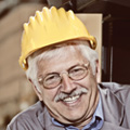 An older man with gray hair and glasses smiles while wearing a yellow construction hard hat and a gray jacket, standing in an industrial setting—just the kind of worker injury compensation lawyers NSW are committed to helping.