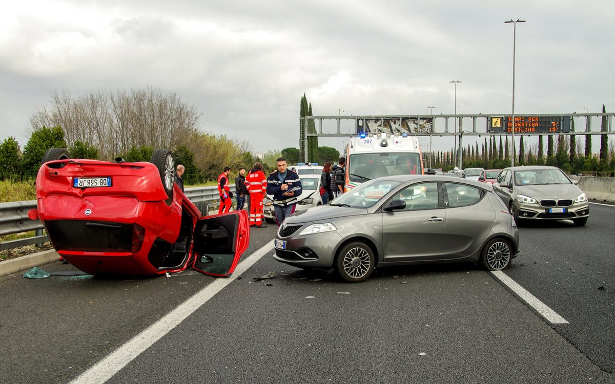 A red car is overturned on a highway next to a silver car with front damage. Emergency responders, an ambulance, and several people are at the scene. Debris is scattered on the road under cloudy skies.