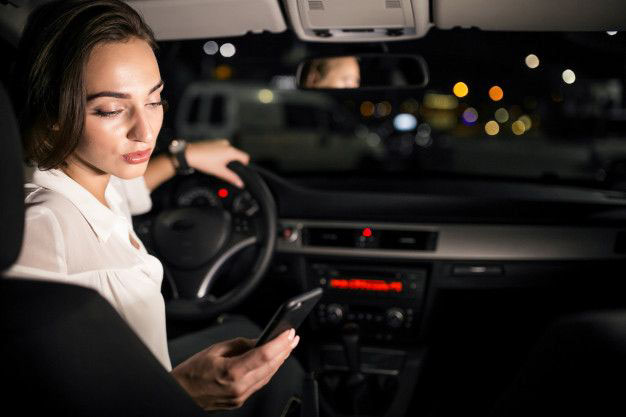 A woman sitting in a car at night looks at her phone while her hand rests on the steering wheel. The car interior is dimly lit, and city lights are visible through the windshield.