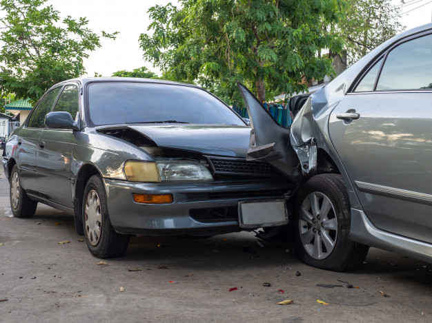 Two cars involved in a rear-end collision; the front of a dark sedan is damaged after crashing into the back of a silver car, which also shows damage. Trees and pavement are visible in the background. Two cars involved in a rear-end collision; the front of a dark sedan is damaged after crashing into the back of a silver car, which also shows damage. Trees and pavement are visible in the background.