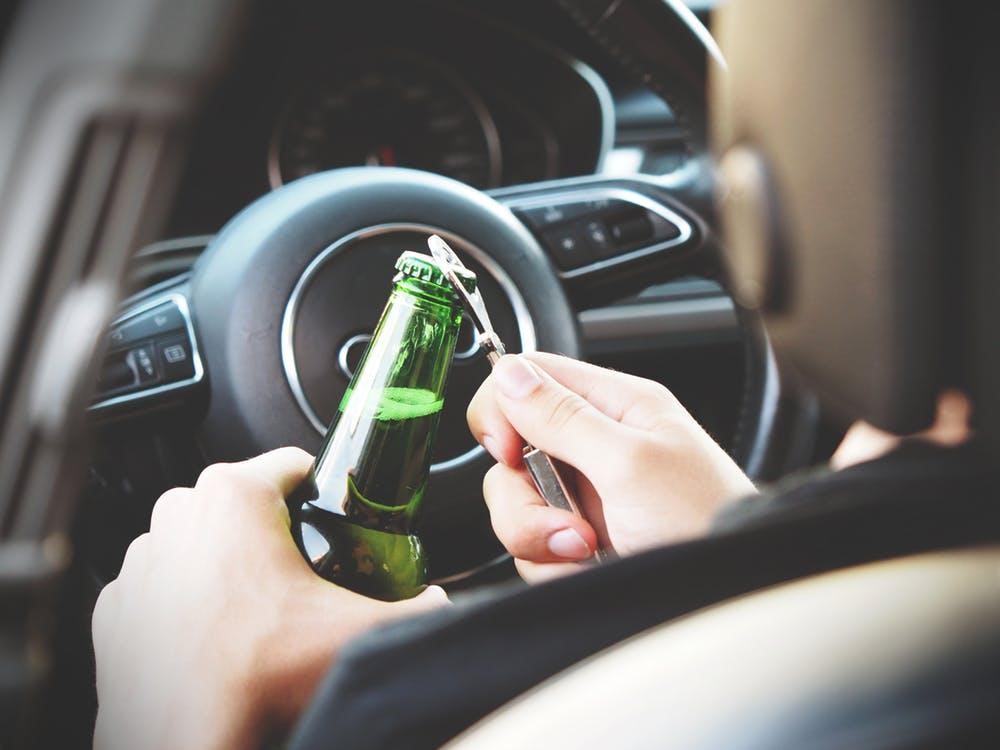 A person sitting in a car uses a bottle opener to open a green beer bottle, holding it near the steering wheel with the dashboard visible in the background.