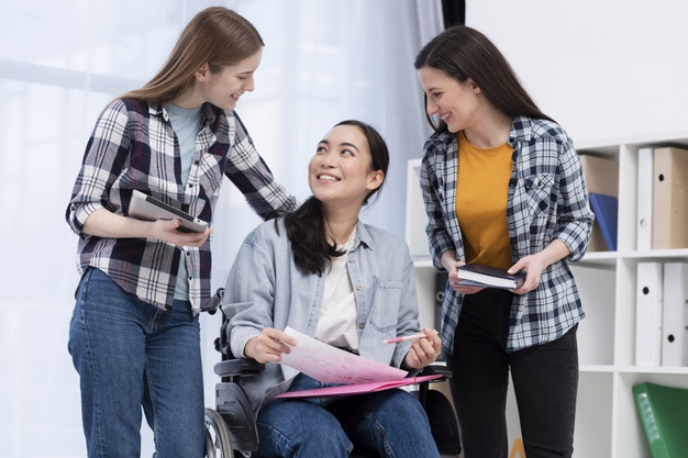 Three young women, one in a wheelchair holding papers, smile and talk together indoors. Two others stand beside her, holding books and a tablet, showing a supportive and friendly atmosphere.
