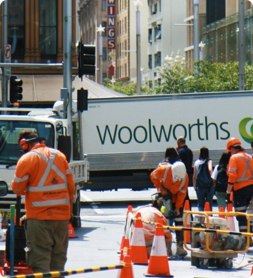 Construction workers in orange safety gear work on a city street near orange cones and machinery, with a Woolworths truck and several pedestrians visible in the background.
