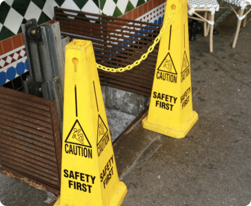 Two yellow "Caution: Safety First" signs and a yellow chain block access to an open stairwell or hole on a sidewalk near a tiled wall, warning pedestrians of a potential hazard.