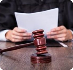 A judge in a black robe holds papers at a desk with a wooden gavel and sound block in the foreground.