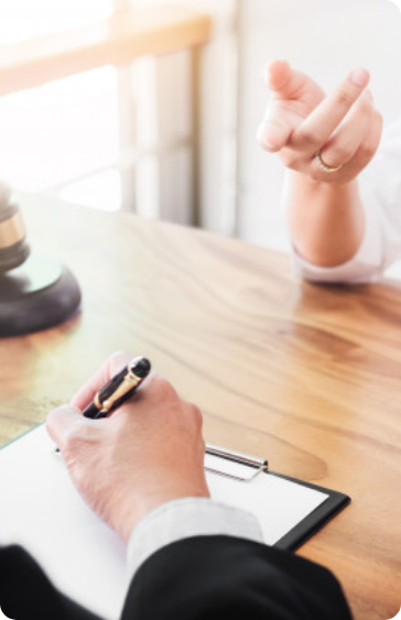 Two people sit at a wooden table; one is writing on a clipboard, while the other gestures with their hand. A gavel is visible on the table, suggesting a legal or professional consultation.