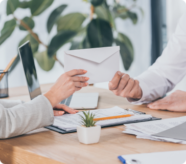 Two people sitting at a desk exchange a white envelope. Documents, a laptop, and a small potted plant are on the desk, with green leaves in the background. Two people sitting at a desk exchange a white envelope. Documents, a laptop, and a small potted plant are on the desk, with green leaves in the background.