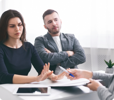 A woman with long brown hair sits at a table, refusing to sign documents offered by a person out of frame, while a man beside her looks on with crossed arms. An electronic tablet and small plant are on the table. A woman with long brown hair sits at a table, refusing to sign documents offered by a person out of frame, while a man beside her looks on with crossed arms. An electronic tablet and small plant are on the table.