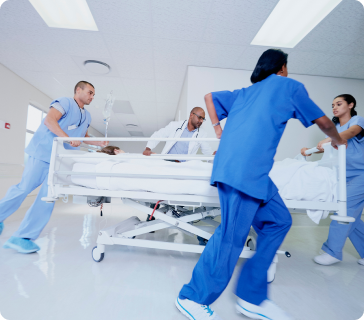 Four medical professionals in blue scrubs and a white coat rush a patient on a hospital bed down a brightly lit hallway, indicating an emergency situation. Four medical professionals in blue scrubs and a white coat rush a patient on a hospital bed down a brightly lit hallway, indicating an emergency situation.