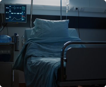 A hospital bed with blue sheets sits empty in a dimly lit room, next to medical equipment and a monitor displaying vital signs. A hospital bed with blue sheets sits empty in a dimly lit room, next to medical equipment and a monitor displaying vital signs.