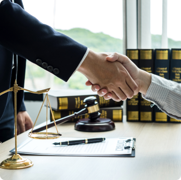 Two people shaking hands across a desk with legal documents, a gavel, scales of justice, and law books, symbolizing a legal agreement or partnership in a law office. Two people shaking hands across a desk with legal documents, a gavel, scales of justice, and law books, symbolizing a legal agreement or partnership in a law office.