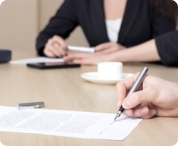 Two people sit at a table during a meeting. One person is writing on a document with a pen, while the other holds a pen and sits near a cup and saucer. Both are dressed in business attire.