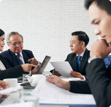 Four business professionals in suits sit at a table with documents, a laptop, and coffee cups, engaged in a serious meeting or discussion in a modern office with a white brick wall background. Four business professionals in suits sit at a table with documents, a laptop, and coffee cups, engaged in a serious meeting or discussion in a modern office with a white brick wall background.