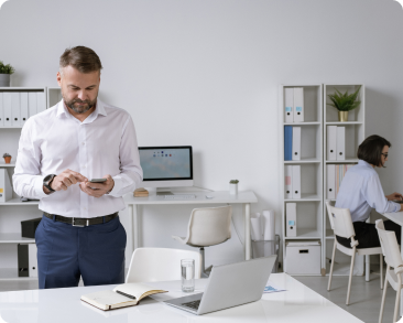 A man in business attire stands at a desk using his phone in a modern office, while a woman works at another desk in the background surrounded by shelves and office supplies. A man in business attire stands at a desk using his phone in a modern office, while a woman works at another desk in the background surrounded by shelves and office supplies.