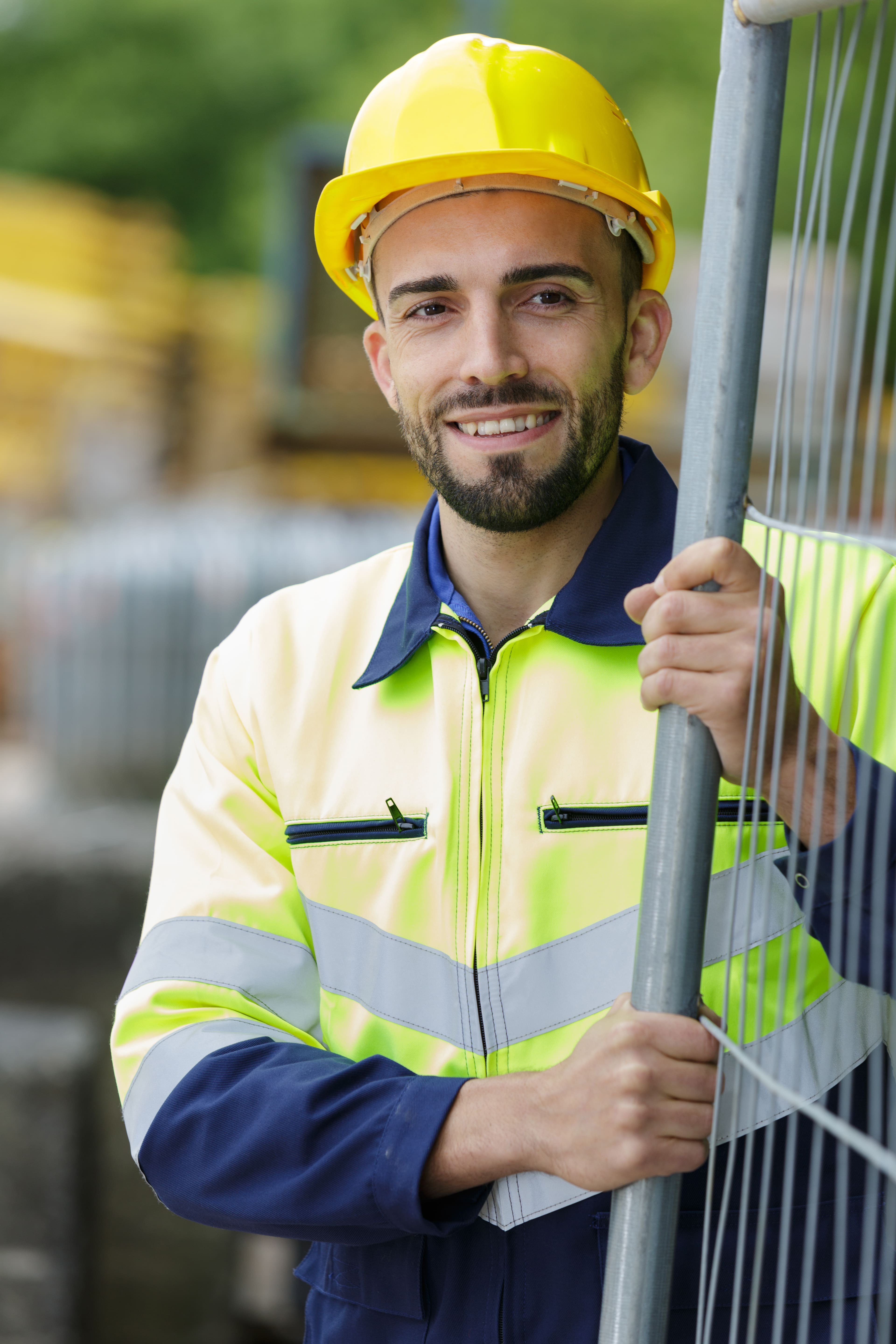 A smiling construction worker wearing a yellow hard hat and high-visibility jacket stands outdoors, holding onto a metal fence. The background is blurred, showing a construction site. A smiling construction worker wearing a yellow hard hat and high-visibility jacket stands outdoors, holding onto a metal fence. The background is blurred, showing a construction site.