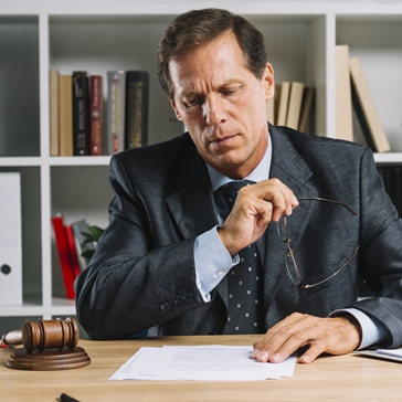 A man in a suit sits at a desk, holding eyeglasses and reviewing documents. A gavel and legal books are on the desk, and shelves with books are in the background, suggesting a legal or office setting.