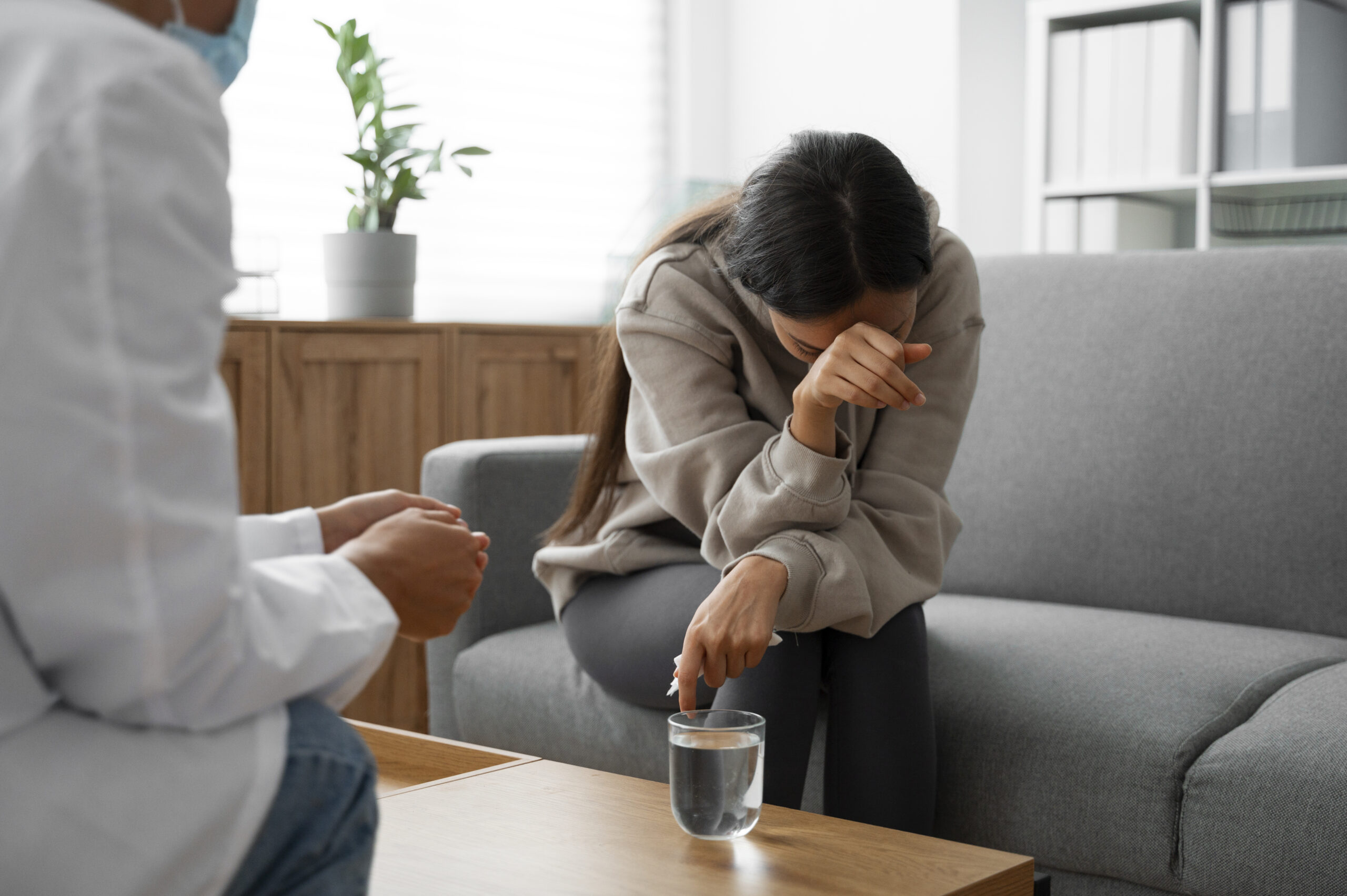 A person sits on a gray couch, visibly upset with their head down and arm covering their face, while another person, likely a professional, sits nearby offering support. A glass of water is on the table in front of them.